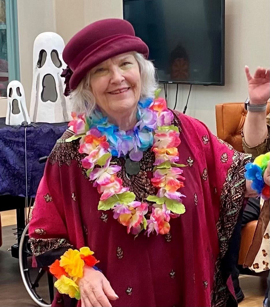 An elderly woman wearing a burgundy hat and colorful floral lei smiles in a festive indoor setting.