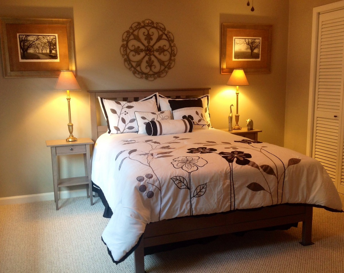 A cozy bedroom featuring a neatly made bed with decorative bedding, accompanied by warm lighting from lamps on side tables and wall art completing the decor.
