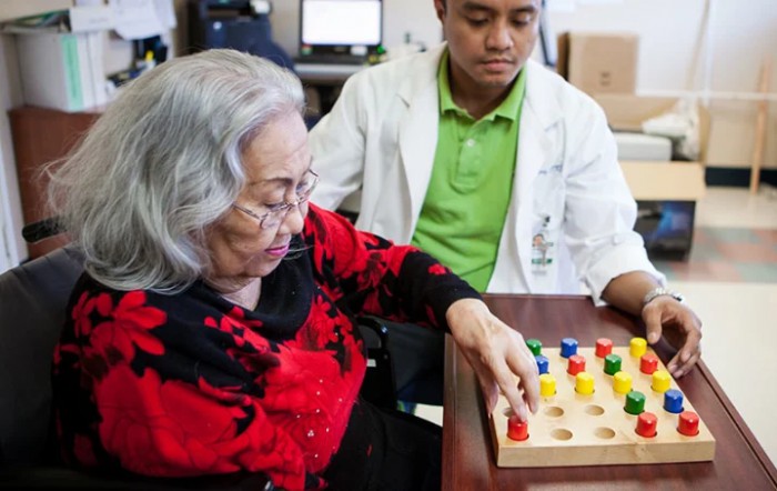 An elderly woman engages in a therapy exercise under the supervision of a healthcare professional.