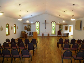Interior view of a chapel featuring stained glass windows and arranged seating with a cross on the wall.