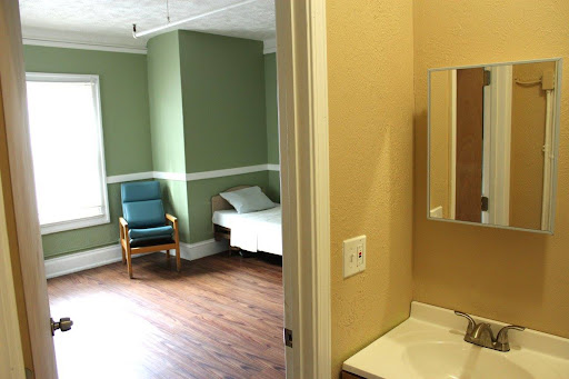 A simple and calm room featuring a bed, chair, and a bathroom area.