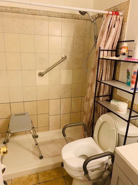 A well-equipped bathroom featuring a toilet with a support rail, a shower area with a bath chair and grab bar, and storage shelves filled with various toiletries.