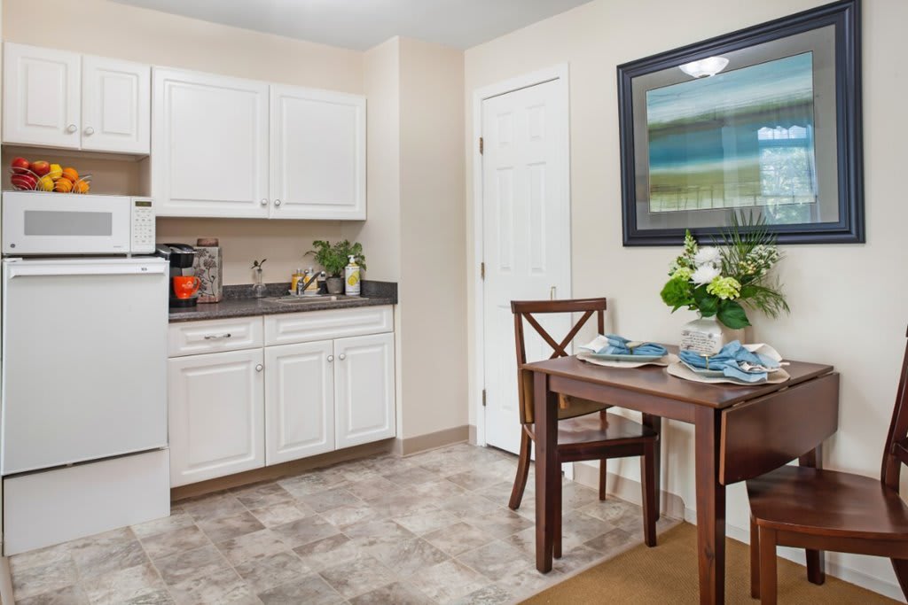 A cozy kitchen and dining area with white cabinets, a small table set for two with flowers, and artwork on the wall.