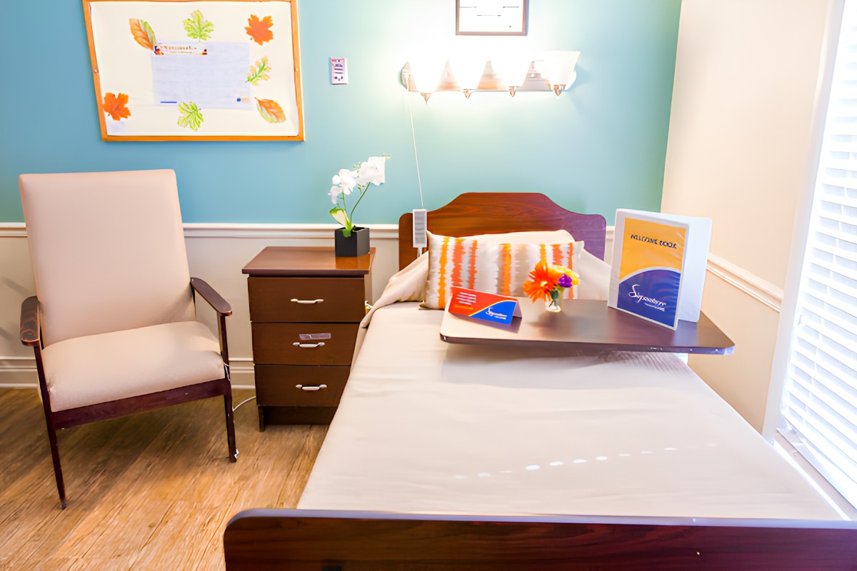 A well-organized nursing home room with a single bed, bedside table, and comfortable chair.