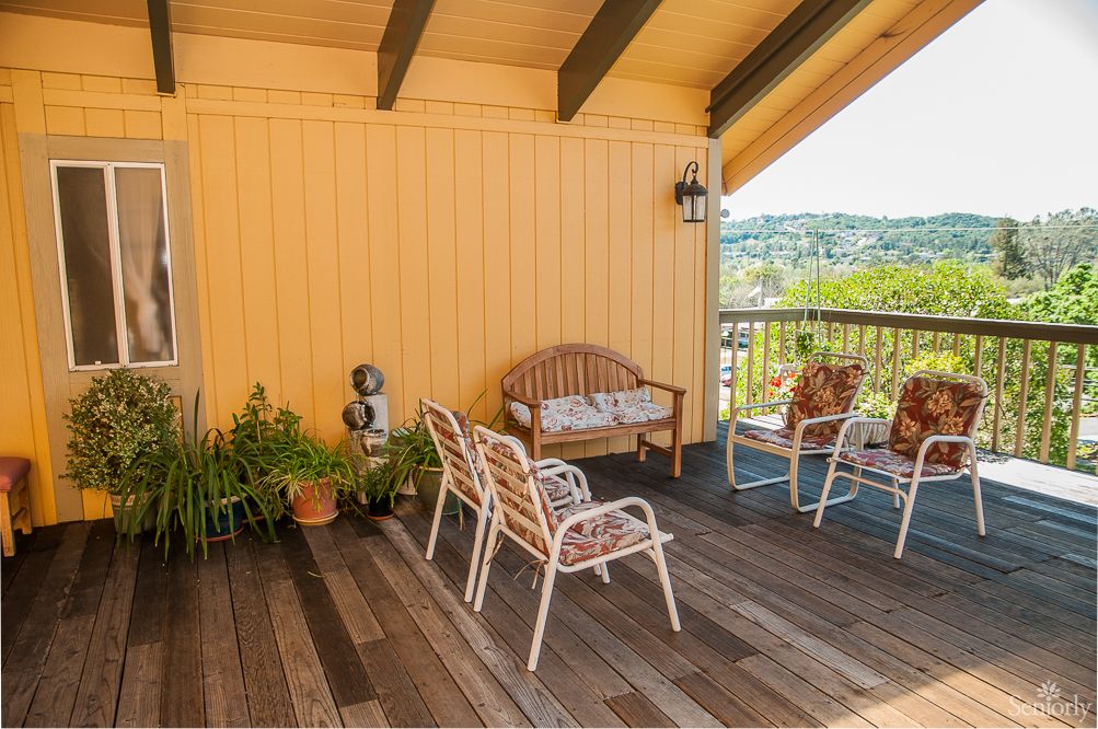 A cozy outdoor patio with wooden flooring, featuring several chairs and a bench surrounded by potted plants.