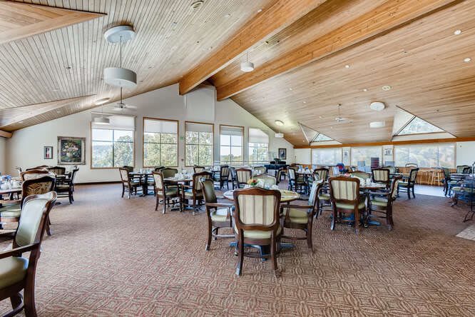A spacious and well-lit dining area with multiple tables and chairs, featuring large windows and a wooden ceiling.