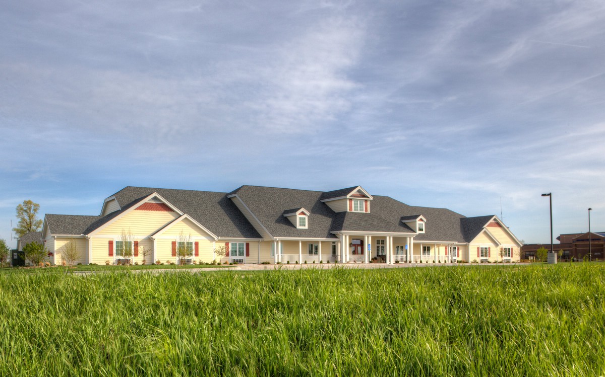 A large farmhouse with a wide grassy field in the foreground and a cloudy sky above.