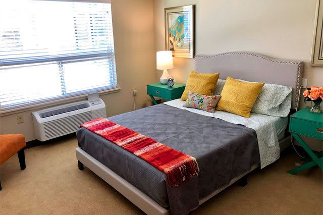 A cozy bedroom with a neatly made bed, featuring colorful pillows and a red-striped throw blanket, next to a teal nightstand and a lamp.