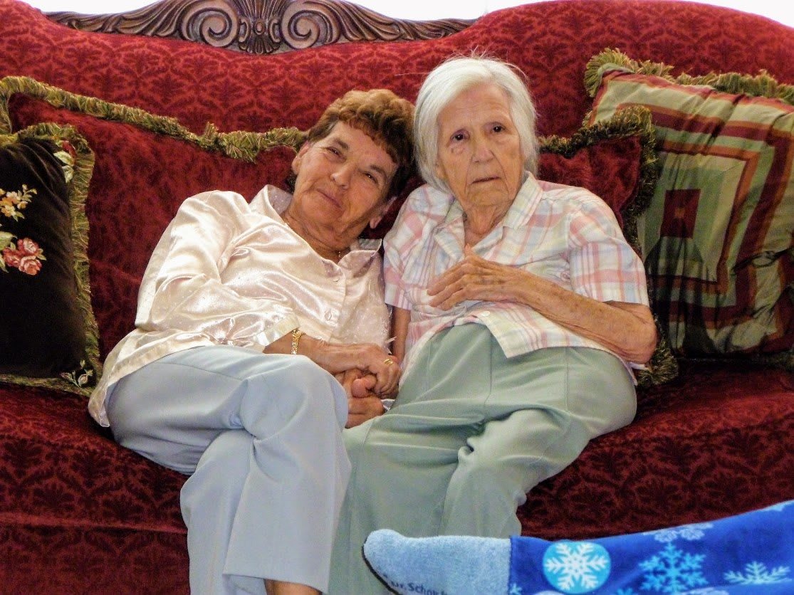 Two elderly women sitting closely together on a couch, sharing a warm moment.
