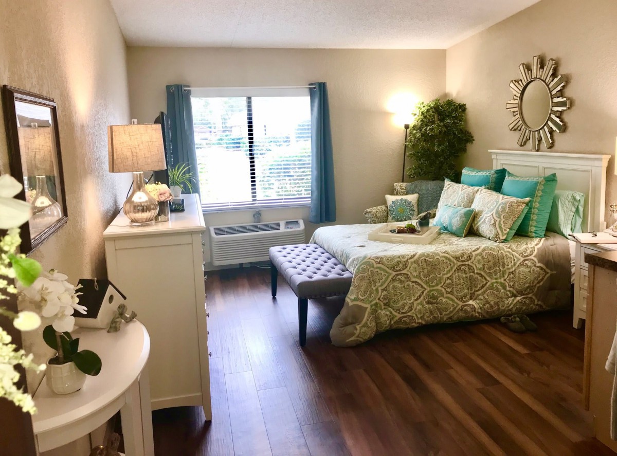 A cozy and well-decorated bedroom with a large window, featuring a bed adorned with green and turquoise bedding, hardwood floors, and decorative plants.