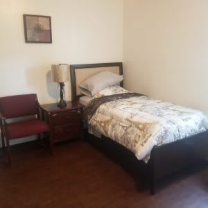 A simple bedroom with a single bed, nightstand, lamp, and a chair on a hardwood floor.