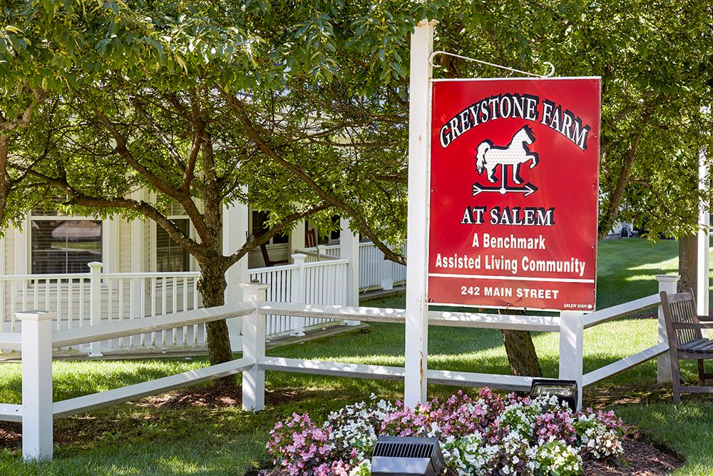 Sign for Graystone Farm at Salem, a senior living community, surrounded by trees and a white fence.