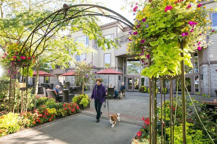 A person walking a dog through a beautifully landscaped garden courtyard with flowers and outdoor seating.