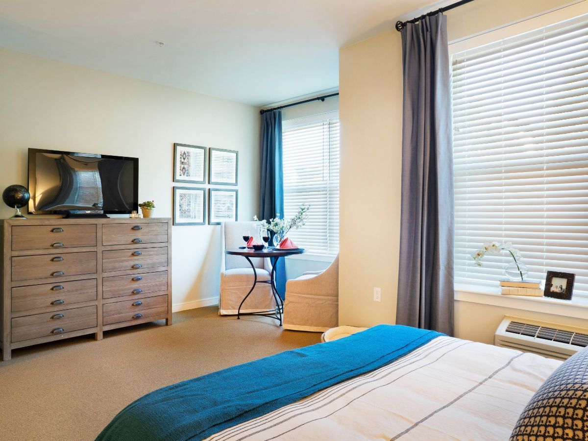A cozy bedroom with modern decor, featuring a dresser with a television and large windows with curtains.