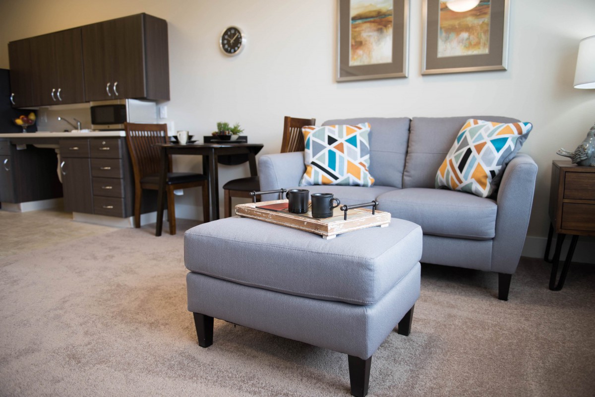 A modern living room with a cozy sofa, colorful pillows, and a stylish ottoman. The space is complemented by a dining area in the background and elegant artwork on the wall.