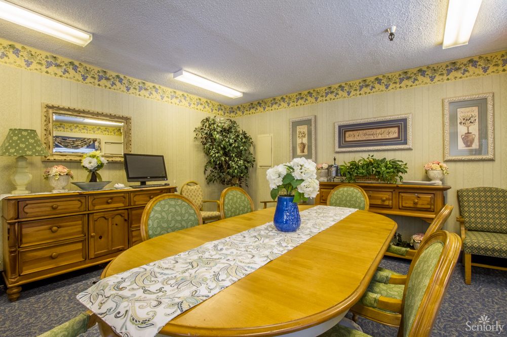 A cozy dining room with a wooden table adorned with a floral centerpiece and surrounded by chairs, complemented by decorative elements in the background.