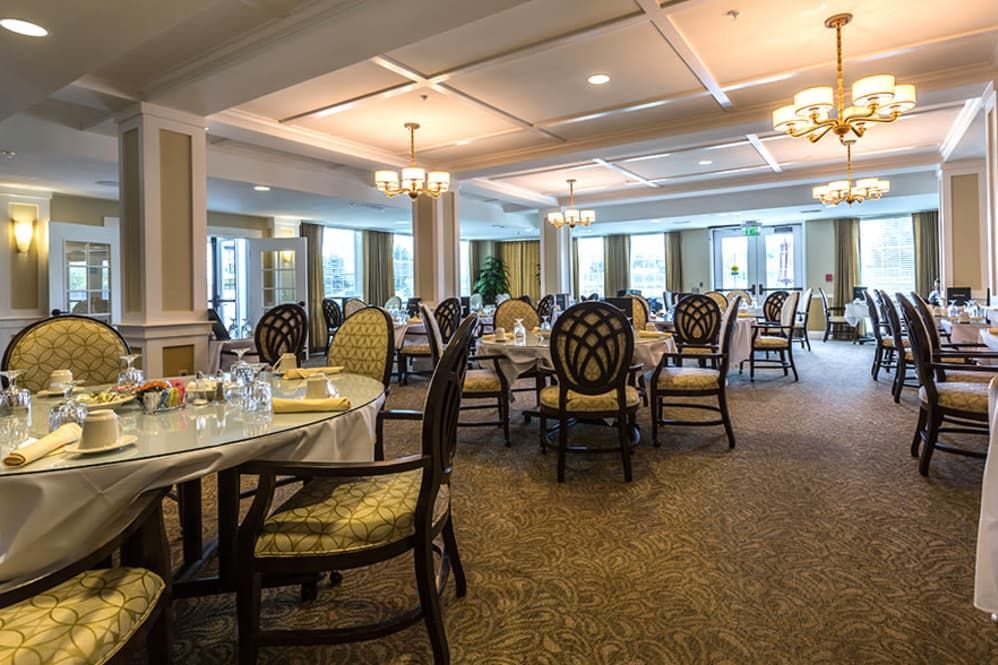 An elegant dining room setup with multiple tables and chairs, featuring chandeliers and large windows.