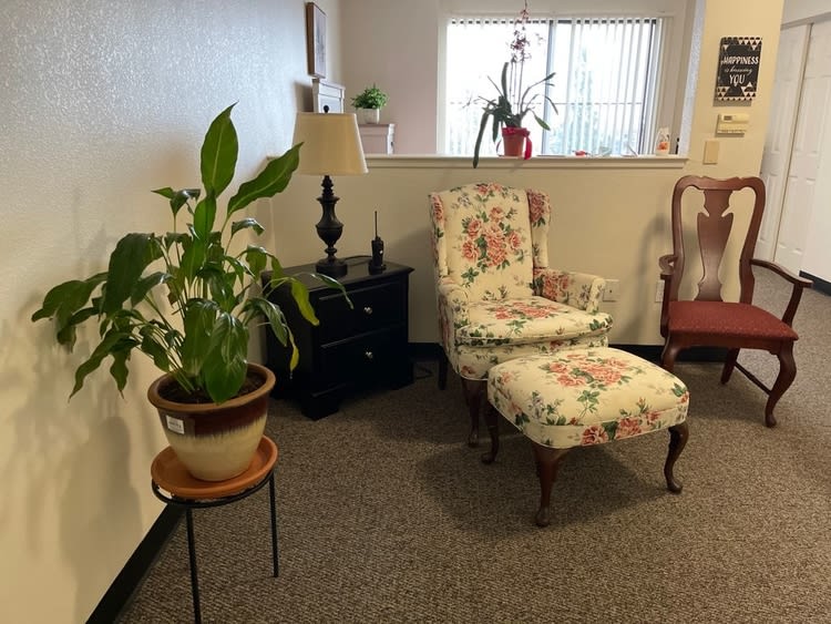 A cozy living room corner featuring a floral patterned chair and ottoman, a lamp on a side table, and indoor plants by the window.