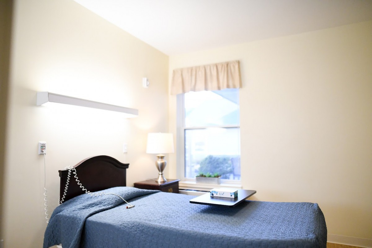 A minimalist bedroom featuring a neatly made bed with a blue cover, a bedside lamp, and a window letting in natural light.