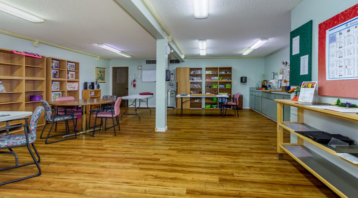 A spacious classroom with wooden floors, tables, chairs, and shelves under bright lighting.