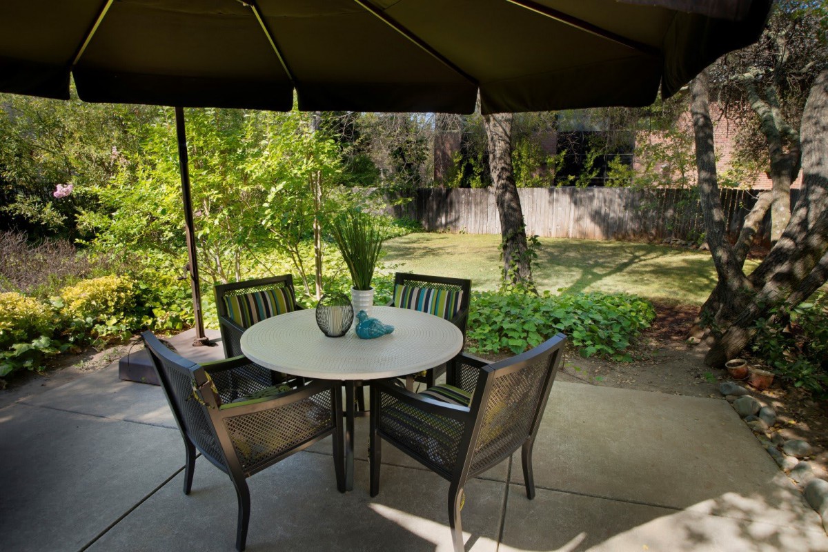 A peaceful outdoor patio with a round table and chairs, shaded by a large umbrella, surrounded by lush greenery.