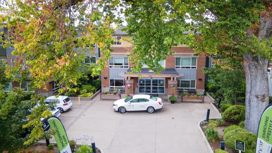 A view of a brick building with cars parked in the driveway, surrounded by trees.