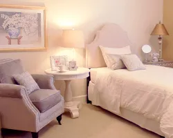 A cozy and elegant bedroom with a soft color palette, featuring a bed, armchair, lamp, and wall art.