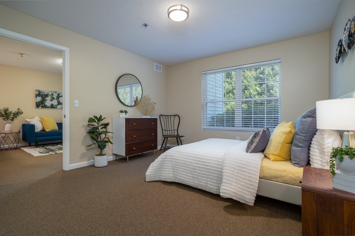 A cozy, minimalist bedroom with a neatly made bed, a chest of drawers with a round mirror above it, and a window letting in natural light.