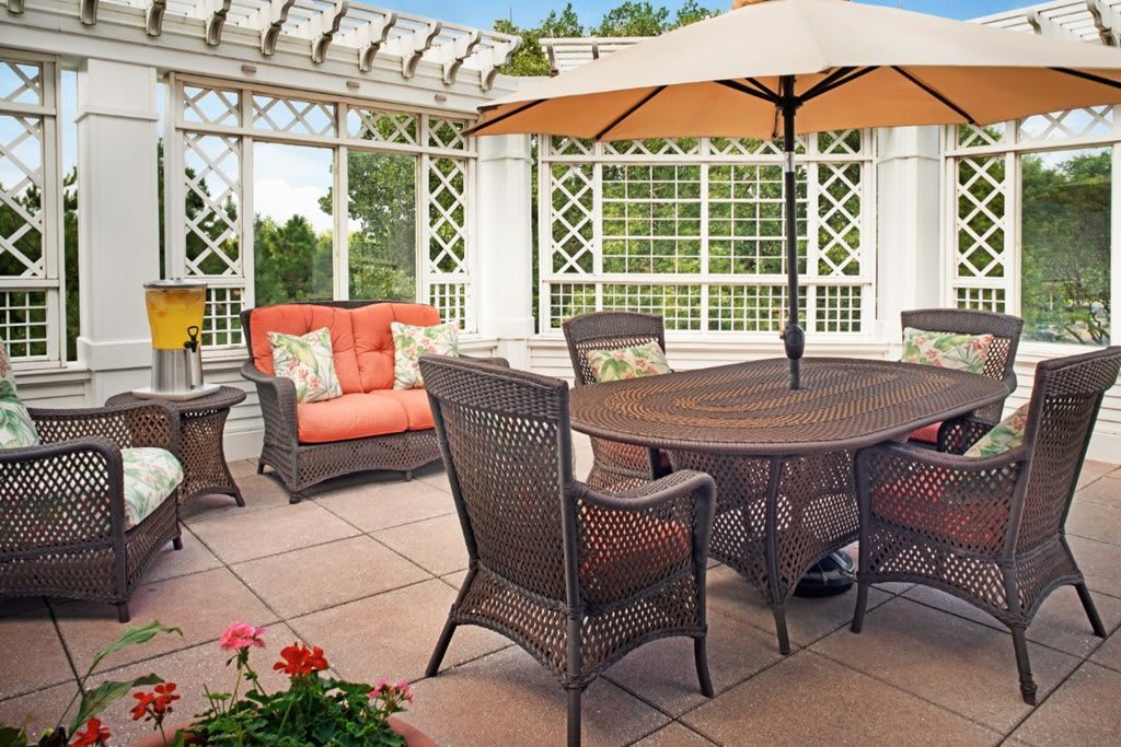 A cozy outdoor patio setup with wicker furniture, a large umbrella, and potted plants under a pergola.