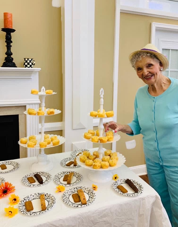 An elderly woman smiling at a dessert table with cupcakes and pastries indoors.