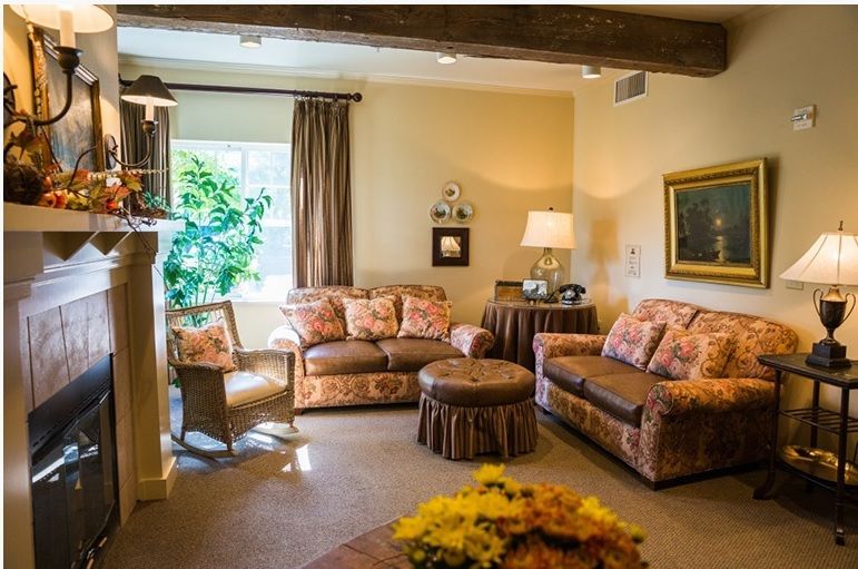 A cozy living room with traditional decor, featuring a sofa set, armchair, and a lamp, illuminated by natural light from a nearby window.