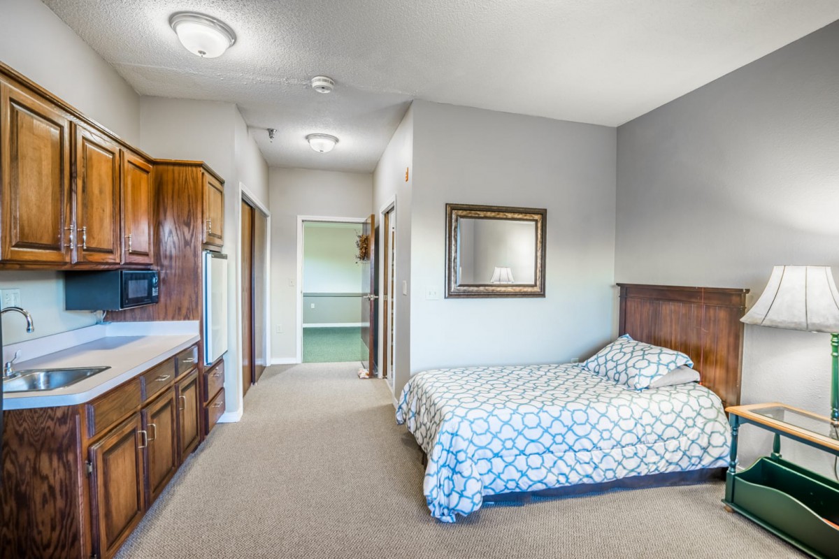 A cozy bedroom featuring a bed with a patterned duvet, a wooden headboard, and a mirror on a light gray wall. The room includes carpeted flooring and leads to a walk-in closet.