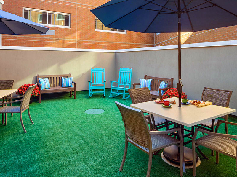 A cozy outdoor patio area featuring a table with chairs, benches with decorative pillows, and umbrellas over green turf.