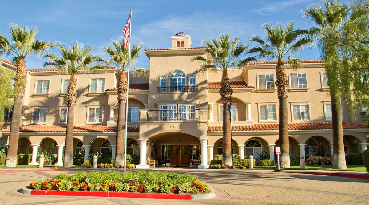 A large building with a grand entrance surrounded by palm trees and neatly arranged flower beds.