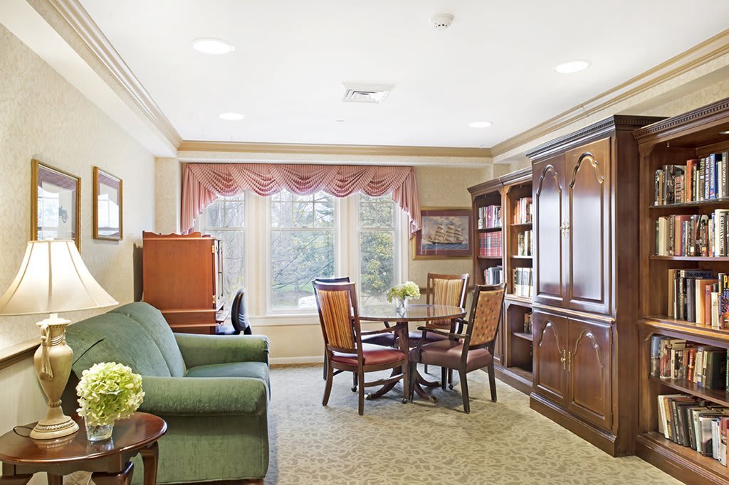 A cozy living room with a sofa, round table and chairs, bookshelves, and a large window with curtains.