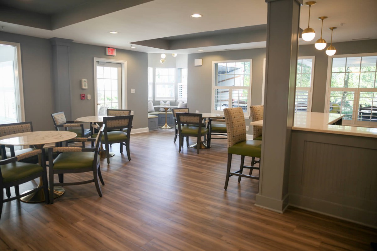 A cozy dining area with tables and chairs, featuring wood flooring and ample natural light from the windows.