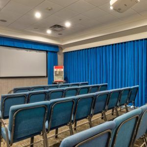 Empty conference room with rows of chairs facing a projector screen and blue curtains.