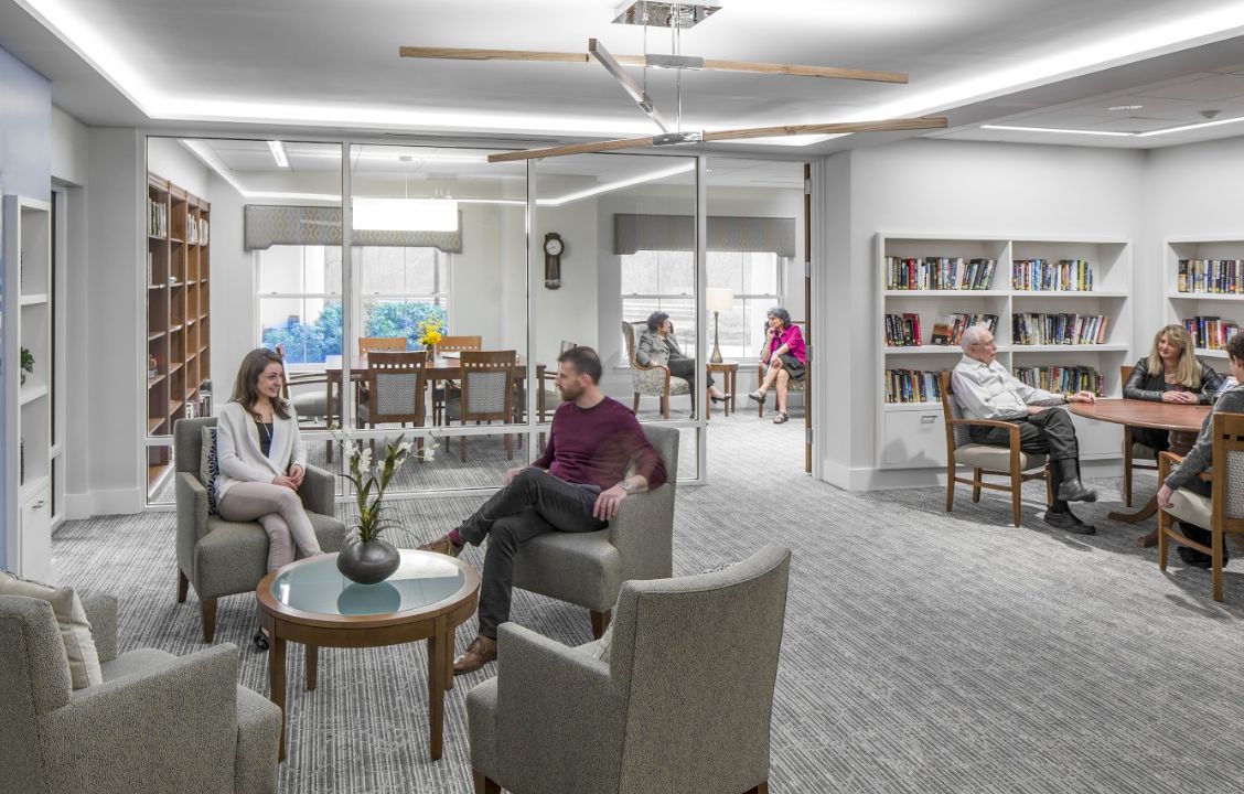 A modern office setting with people engaging in conversation and working in a comfortable seating area with glass walls and bookshelves.