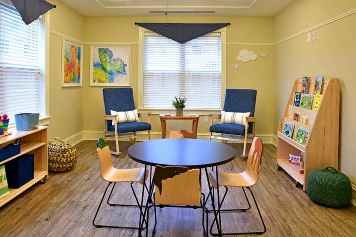 A cozy children's playroom with a round table, wooden chairs, and a bookshelf filled with books and games.