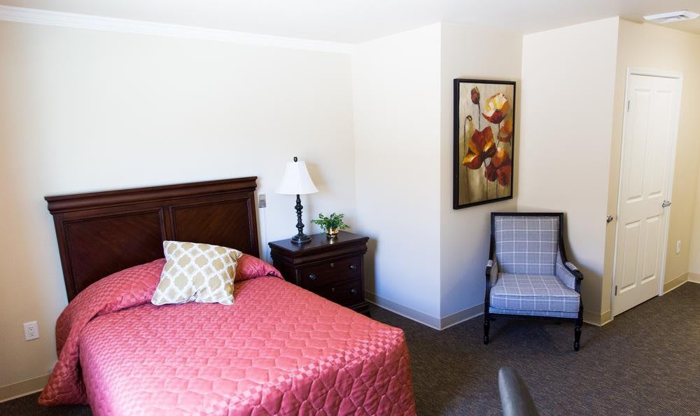 A neat bedroom with a red bedspread, nightstand with lamp, and decorative painting.