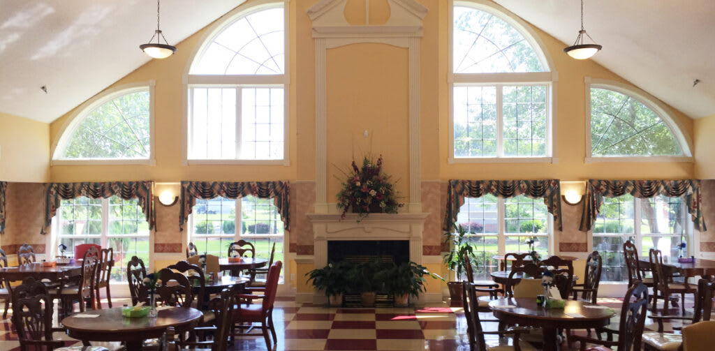 A bright dining room with large windows and classic decor, featuring a fireplace surrounded by tables and chairs.