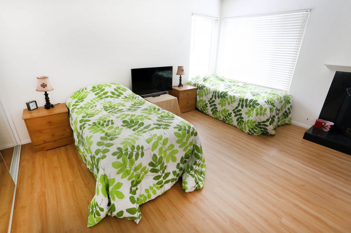 A bright bedroom with two twin beds featuring green bedding and a television on a wooden floor.