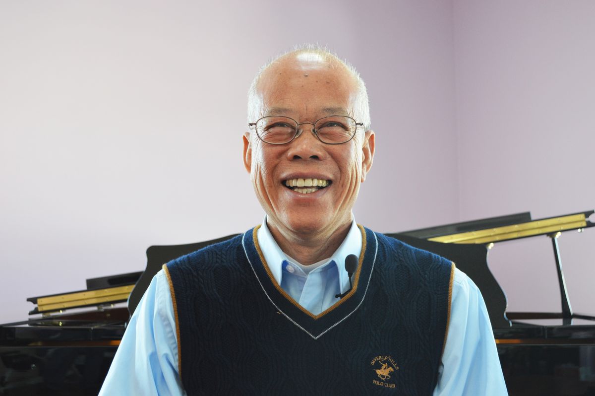 An elderly man wearing glasses and a sweater vest, smiling warmly indoors.