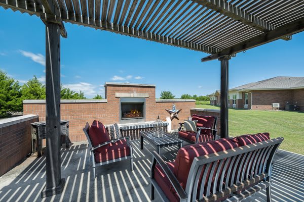 A sunny outdoor patio with a pergola, furnished with striped chairs and a central fireplace.