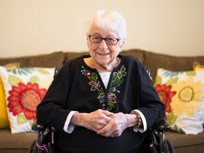 An elderly woman wearing glasses, smiling while seated indoors with floral pillows in the background.