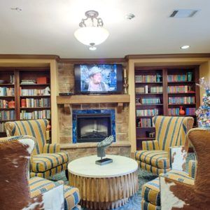 A cozy library room with bookshelves, a fireplace, and comfortable seating around a central table.