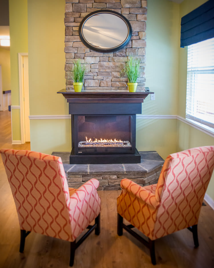 A cozy living room setup featuring two patterned chairs facing a stone fireplace, adorned with plants and a mirror.