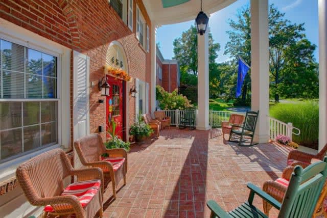 A charming front porch with brick flooring, adorned with chairs and plants, featuring large columns and a hanging lantern.