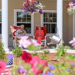 A cozy porch with people relaxing in chairs surrounded by vibrant flowers and hanging plants.