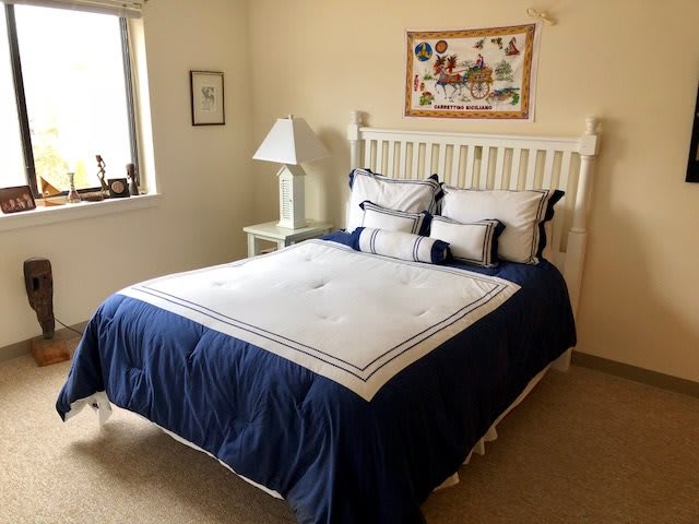 A cozy bedroom featuring a neatly made bed with blue and white bedding, a bedside lamp, artwork on the wall, and natural light from a window.